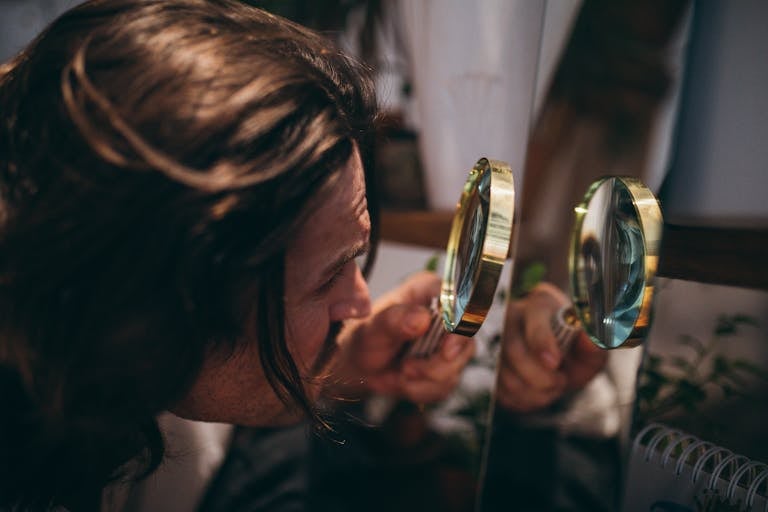 Close-up shot of a man with long hair using a magnifying glass to inspect his reflection in a mirror.